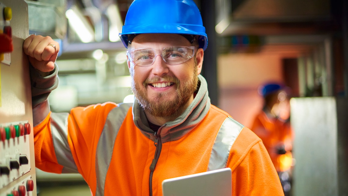 An industrial service engineer smiles to camera before he conducts a safety check of a control panel in a boiler room. He is wearing hi vis, hard hat, safety glasses and holding a digital tablet as he conducts a safety inspection.