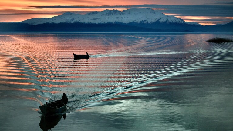 Two fishing boats on a lake in front of a mountain panorama at dusk.